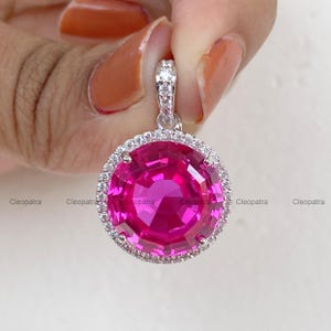 May include: A round pendant featuring a large, faceted, vibrant pink gemstone at its center. The stone is encircled by a halo of small, clear, sparkling white stones set in a silver-toned metal frame, with a matching stone-encrusted bail.