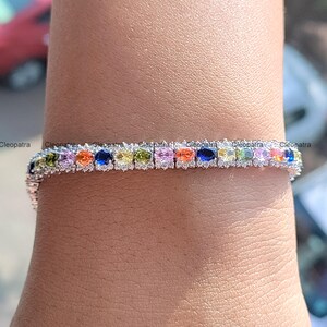 May include: A silver bracelet with a row of colorful oval-cut gemstones. The gemstones include shades of blue, green, yellow, orange, pink, and purple. The bracelet is set with small, clear stones around each colored gem. The word "Cleopatra" is visible.
