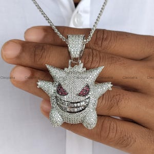 Puede incluir: Un colgante plateado con forma de criatura de dibujos animados, totalmente incrustado con pequeñas piedras claras. La criatura presenta dos orejas puntiagudas, dos ojos de piedra roja y una amplia sonrisa con piedras de corte baguette.
