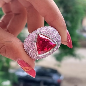 Pode incluir: Um anel com uma grande pedra central triangular de cor vermelho-profundo, montada numa banda de metal prateado. Toda a superfície da banda está densamente incrustada com numerosas pedras pequenas, redondas e cor-de-rosa claro, criando um aspeto texturado e cintilante.