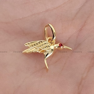 May include: A gold-coloured hummingbird pendant with a red gemstone eye. The pendant has detailed wing and body designs, and a loop for attaching to a chain. The hummingbird is a symbol of joy and beauty.