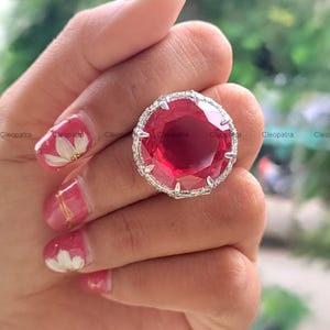 May include: A large, round, faceted red gemstone ring set in a silver-toned metal band. The setting features a decorative border of small, clear, sparkling stones surrounding the central red stone, held in place by multiple silver prongs.