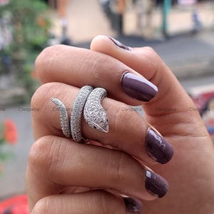 Puede incluir: Un anillo de serpiente plateado con un diseño texturizado y tachonado de gemas. La cabeza de la serpiente es puntiaguda y el anillo se envuelve alrededor del dedo. El anillo se muestra en una mano con esmalte de uñas morado oscuro.