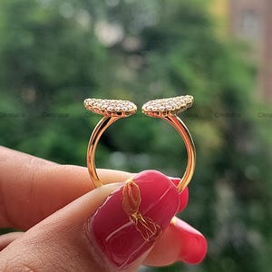 May include: A gold-colored ring with two open, leaf-shaped designs encrusted with small, clear gemstones. The ring is held up against a blurred green background. The ring's design is open, with the two gemstone-covered sections facing outward.
