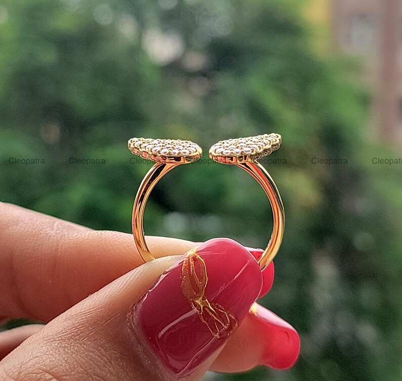 May include: A gold-colored ring with two open, leaf-shaped designs encrusted with small, clear gemstones. The ring is held up against a blurred green background. The ring's design is open, with the two gemstone-covered sections facing outward.