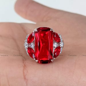 Puede incluir: Un anillo plateado con una gran piedra roja rectangular facetada como pieza central. El anillo está adornado con piedras rojas más pequeñas en forma de marquesa y piedras de acento redondas y transparentes.