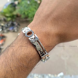 Könnte beinhalten: Ein silberfarbenes Armband mit geometrischem Design. Das Armband hat rechteckige und kreisförmige Glieder mit einem Verschluss. Das Armband wird am Handgelenk getragen.