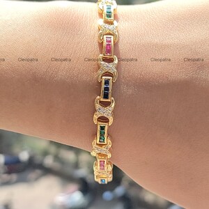 May include: A gold bracelet with a repeating pattern of X's and small clear stones. The bracelet is accented with rows of colorful stones, including green, blue, red, and pink.