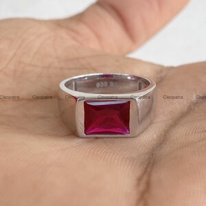 May include: A silver-toned ring featuring a rectangular, faceted, deep pinkish-red gemstone set in the center. The interior of the band is clearly stamped with the text 935 SIL, indicating the metal composition of the polished, smooth ring band.