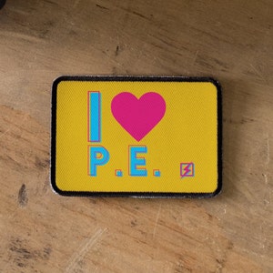 May include: A yellow patch with black stitching features the text "I ♥ P.E." in blue and pink lettering. A pink heart is in the centre of the patch. A lightning bolt symbol is in the bottom right corner.