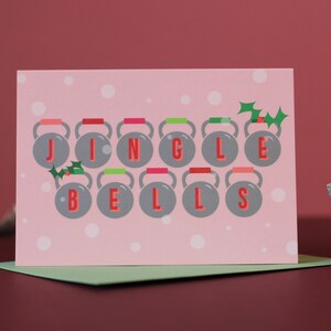 May include: A pink Christmas card with the words "JINGLE BELLS" spelled out using kettlebells. The kettlebells are gray with red, green, and pink accents. Holly sprigs are placed above the letters. The background is light pink with white polka dots.