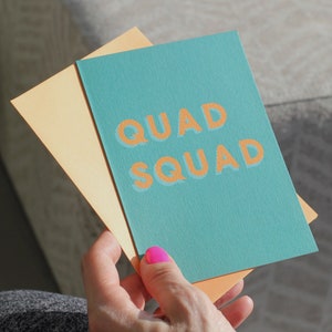 May include: A teal card with the text "QUAD SQUAD" in orange letters. The card is folded in half and held by a hand with pink nail polish.