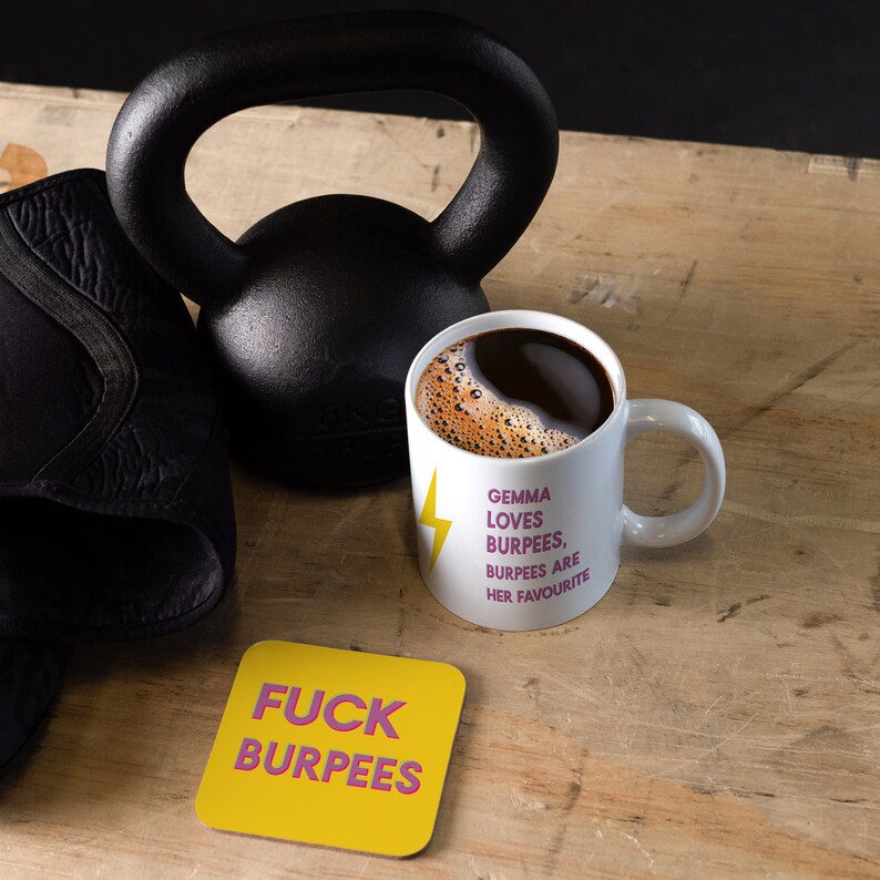 May include: White coffee mug with the text "Gemma loves burpees, burpees are her favourite" in pink lettering and a yellow lightning bolt. A yellow coaster with the text "F*ck burpees" in pink lettering is in the foreground. A black kettlebell and a black workout mat are in the background.