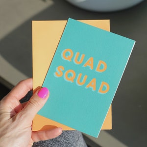 May include: A teal card with the text "QUAD SQUAD" in orange letters. The card is on top of a yellow card.