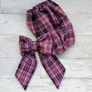 May include: A purple plaid fabric bow and matching scrunchie. The bow has long tails and a large knot. The scrunchie is gathered and elasticized. The plaid pattern includes black, white, and pink lines.