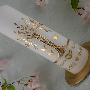 Puede incluir: Vela blanca con un diseño de árbol dorado y mariposas. La vela tiene los nombres "Barbara" y "Josef" y las fechas "06.05" y "15.1967" escritas en ella. La vela está sobre una base dorada.