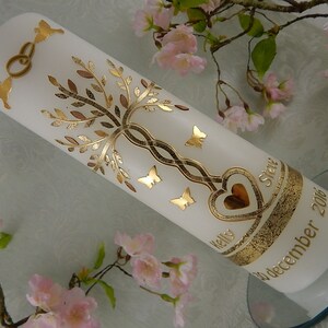 Könnte beinhalten: Weiße Hochzeitskerze mit goldenen Akzenten. Die Kerze zeigt einen Baum mit ineinander verschlungenen Ästen, Schmetterlingen und einem Herzen mit den Worten "Kelly" und "Steve" auf beiden Seiten. Das Datum "10. Dezember 2016" ist auf der Unterseite der Kerze geschrieben.
