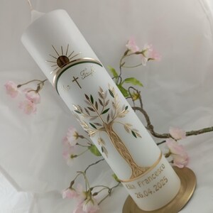 May include: White pillar candle featuring a gold and green tree design, sunburst, and text. The candle has a gold band with the words "Zur Taufe" and a cross. The base has the name "Ella Francesco" and the date "26.04.2025".