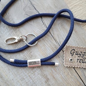 Könnte beinhalten: Ein dunkelblaues Lanyard mit einem silbernen Verschluss, einem silbernen Ring und einem silbernen Anhänger mit dem Wort "MOIN" und einem Anker-Symbol. Ein kleines braunes Etikett mit dem Text "Gugga rella" ist ebenfalls sichtbar.