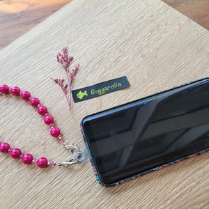 Könnte beinhalten: Ein rosa Perlen-Handyband mit einem silbernen Verschluss, der an einer schwarzen Handyhülle mit einem bunten Muster befestigt ist. Die Hülle trägt den Text "Guggarellia" auf einem schwarzen Etikett.