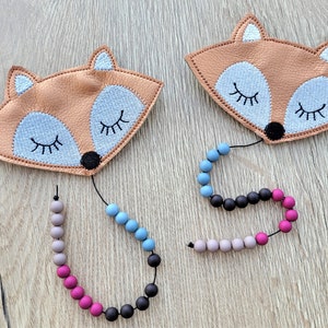 Könnte beinhalten: Zwei fuchsförmige Stofftiere mit gestickten Augen und einer schwarzen Nase. Jedes Spielzeug ist an einer Schnur mit bunten Holzperlen befestigt, darunter rosa, blaue, graue und schwarze Perlen.