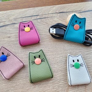 Könnte beinhalten: Eine Sammlung farbenfroher Kabelorganisatoren in Katzenform. Die Organizer sind in verschiedenen Farben erhältlich, darunter Rosa, Türkis, Grün und Weiß. Jede Katze hat eine Knopf-Nase und gestickte Details. Ein schwarzes USB-Kabel wird von einem türkisfarbenen Katzen-Organizer gehalten.