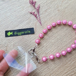Könnte beinhalten: Ein rosa Perlenarmband mit einem silbernen Verschluss. Das Armband besteht aus kleinen, runden Perlen mit einem Perlmutt-Finish. Das Armband liegt auf einer Holzoberfläche mit einem kleinen grün-schwarzen Etikett, auf dem "Guggarella" steht.