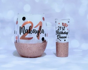 May include: A set of two glasses with rose gold glitter accents. The larger glass has the number 21 and the name Makayla written on it. The smaller glass has the text "21st Birthday Queen" written on it.