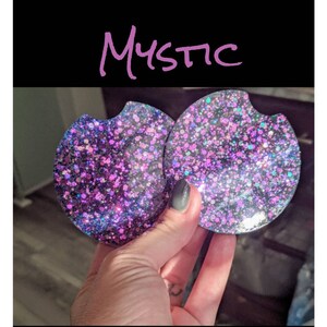 May include: Two round car coasters with a glittery design in shades of purple, pink, and blue. The word "Mystic" is written in cursive above the coasters. The coasters have a small indentation for easy removal from a car cup holder.