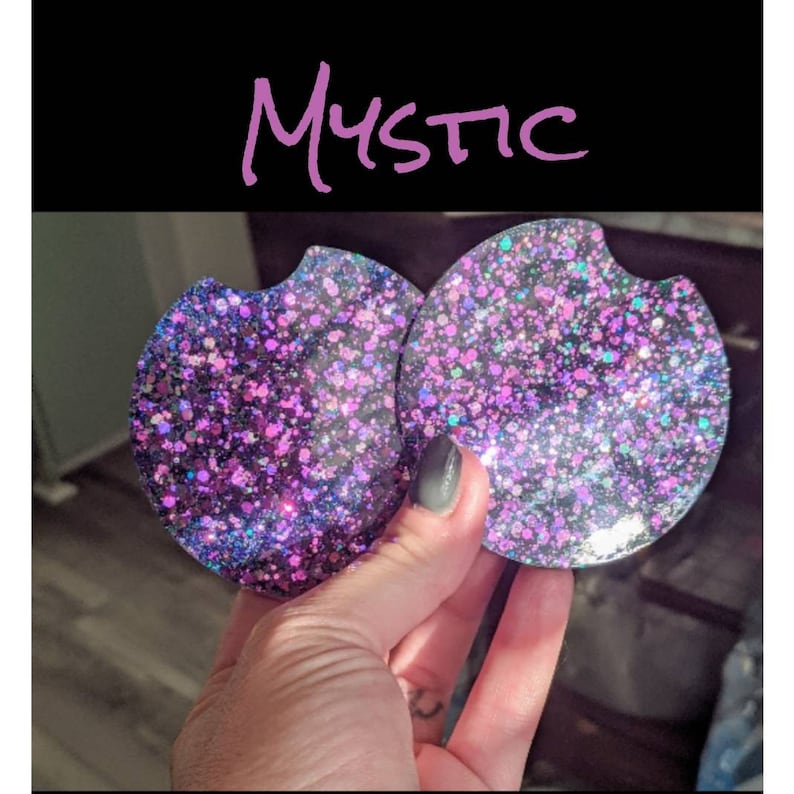 May include: Two round car coasters with a glittery design in shades of purple, pink, and blue. The word "Mystic" is written in cursive above the coasters. The coasters have a small indentation for easy removal from a car cup holder.
