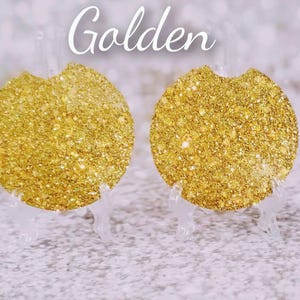 May include: Two round, golden glitter car coasters. The coasters are displayed on clear stands against a blurred background. The word "Golden" is written in cursive across the top of the image.
