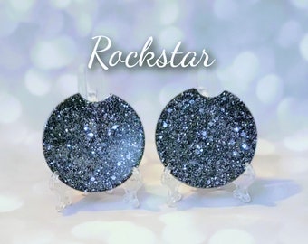 May include: Two round black glitter coasters with the word "Rockstar" printed above them. The coasters are on small clear stands.