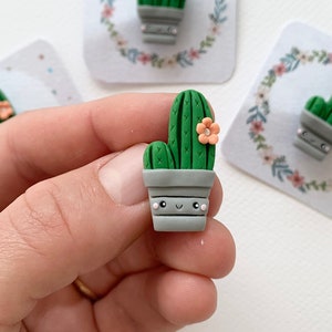 Könnte beinhalten: Drei grüne und graue Kaktus-Figuren aus Ton mit einer rosa Blume obenauf. Der Kaktus steht in einem grauen Topf mit einem fröhlichen Gesicht. Die Figuren stehen auf einem weißen Hintergrund.