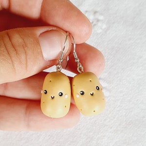 Op de afbeelding: Een paar oorhangers met een schattig cartoon-aardappelontwerp. De aardappelen zijn geel met witte accenten en hebben een vrolijk gezicht.