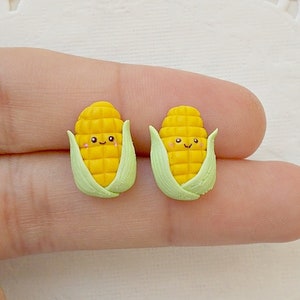 Könnte beinhalten: Ein Paar gelbe Mais-Ohrringe mit grünen Hüllen und lächelnden Gesichtern. Die Ohrringe sind aus Polymer Clay gefertigt und etwa 2,5 cm lang.