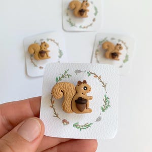 May include: A collection of squirrel-themed pins. Each pin features a brown squirrel holding a dark brown acorn, set against a white square card with a decorative green and brown wreath. The squirrel has a light brown body and a fluffy tail.