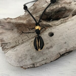 Puede incluir: Un collar de cordón de cuero negro con una cuenta dorada y un colgante de concha de cauri negro.