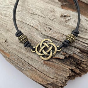Könnte beinhalten: Ein Armband aus schwarzem Lederband mit einem goldfarbenen keltischen Knoten-Anhänger und zwei goldfarbenen Perlenakzenten.