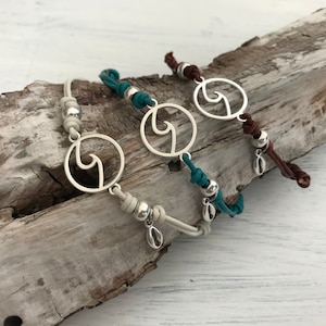Op de afbeelding: Drie armbanden met een zilveren golfbedel, elk met een andere koordkleur: wit, turquoise en bruin.
