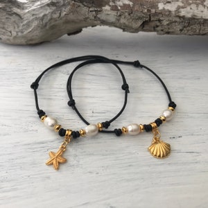 Op de afbeelding: Twee zwarte koordarmbanden met gouden accenten. De ene armband heeft een gouden zeester-bedel en de andere een gouden schelp-bedel. Beide armbanden hebben witte parels.