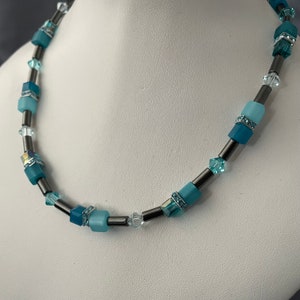 Può includere: Una collana con un mix di perline quadrate blu chiaro e scuro, perline di cristallo trasparente e tubi di metallo color argento. La collana è esposta su un busto di manichino bianco.