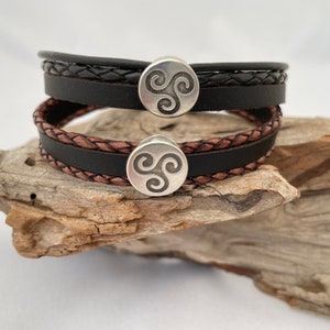 Può includere: Due braccialetti in pelle con un design intrecciato e un ciondolo in metallo argentato con un nodo celtico. I braccialetti sono impilati uno sopra l'altro su un pezzo di legno di deriva.