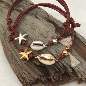Op de afbeelding: Twee bruine leren armbanden met gouden en zilveren zeesterren en schelpbedels. De armbanden hebben kwastjes aan de uiteinden.