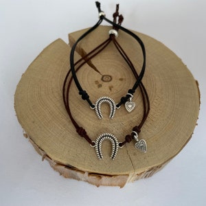 May include: Two leather bracelets with silver horseshoe charms and heart charms. The bracelets are brown and black and are displayed on a wooden surface.
