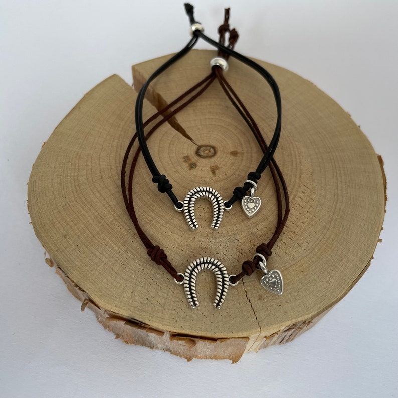 May include: Two leather bracelets with silver horseshoe charms and heart charms. The bracelets are brown and black and are displayed on a wooden surface.