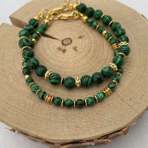 Può includere: Due braccialetti di perline di malachite verde con dettagli dorati. I braccialetti sono sovrapposti su una superficie di legno.