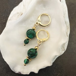Op de afbeelding: Een paar gouden oorringen met groene malachietstenen kralen. De oorringen hebben een hoepelontwerp en zijn versierd met kleinere gouden kralen.