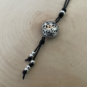 Könnte beinhalten: Eine Halskette aus schwarzem Lederband mit einem silbernen Anhänger, der ein kreisförmiges, filigranes Design aufweist. Der Anhänger ist mit einem Knoten am Band befestigt und das Band hat silberne Perlen.