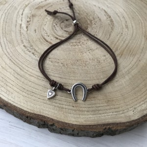 May include: A brown leather cord bracelet with a silver horseshoe charm and a small silver heart charm.