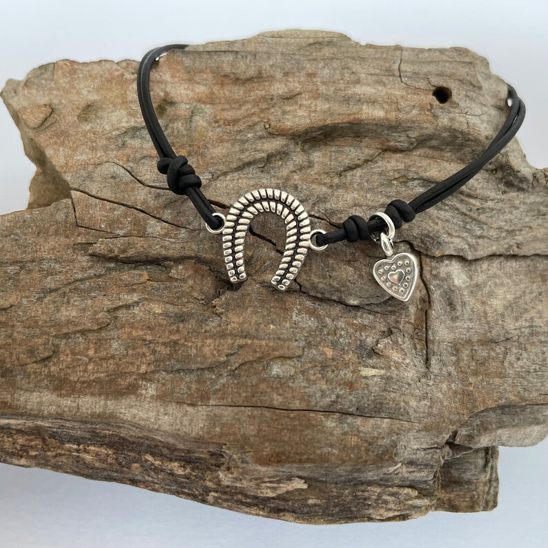 May include: A black leather bracelet with a silver horseshoe charm and a silver heart charm.